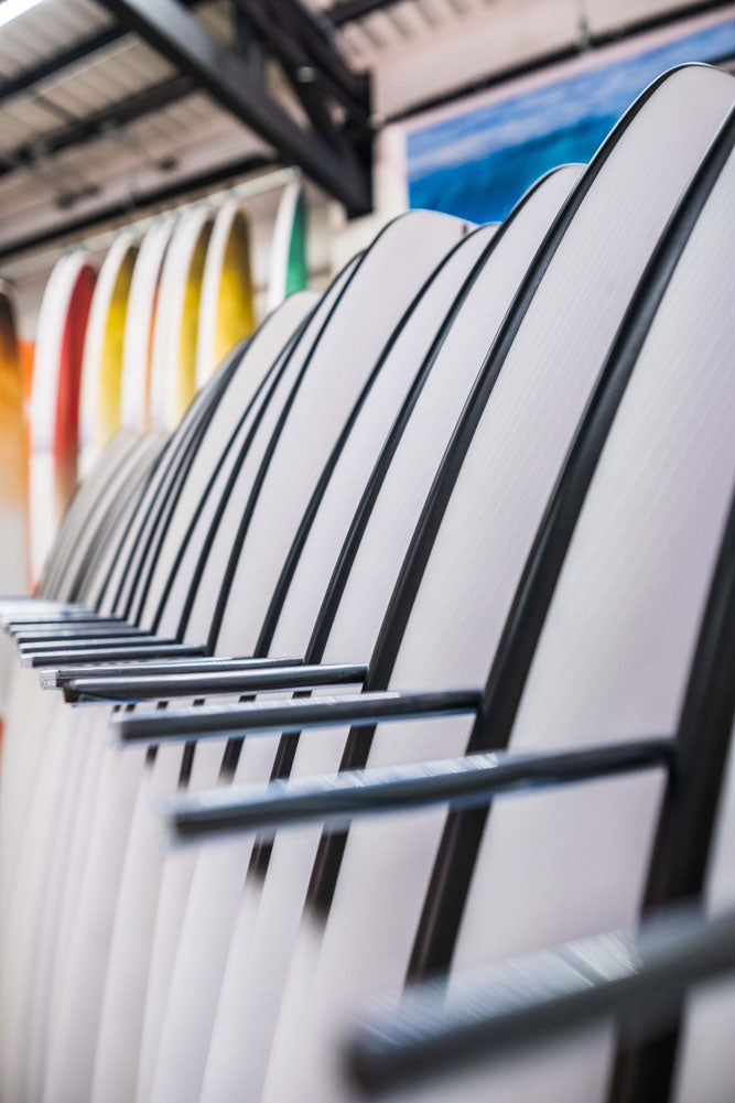Row of white surfboards with black fins on a rack in a warehouse setting.